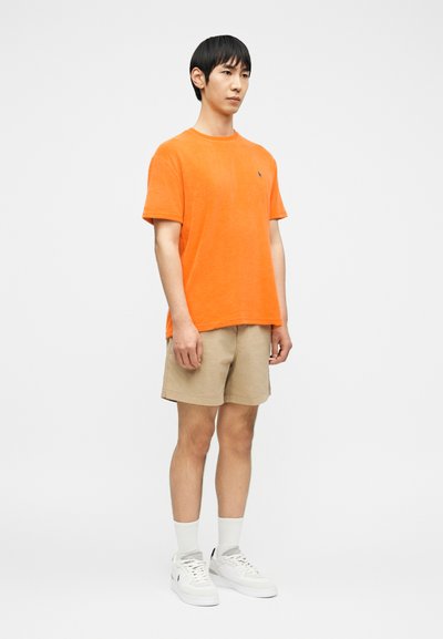 Νεαρός άνδρας που στέκεται φορώντας πορτοκαλί μπλουζάκι με κοντό μανίκι, μπεζ σορτς, λευκές κάλτσες και λευκά αθλητικά παπούτσια σε απλό φόντο.