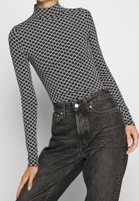 Body a maniche lunghe bianco e nero con colletto alto, caratterizzato da un motivo a zigzag, abbinato a jeans neri a vita alta.