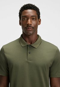 Khaki polo z teksturowanym wzorem, z kołnierzykiem i zapięciem na dwa guziki. Krótkie rękawy i dopasowany krój.