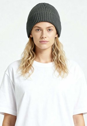 Jonge vrouw met lang blond golvend haar, een grijze geribbelde gebreide muts en een eenvoudige witte oversized T-shirt, recht vooruit kijkend tegen een witte achtergrond.