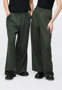 Πράσινο παντελόνι με φαρδιά μπατζάκια και πιέτες, κατασκευασμένο από λείο ύφασμα. Διαθέτει βαθιές τσέπες και συνδυάζεται με μαύρα chunky παπούτσια.