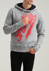 Grauer Hoodie mit einem roten Grafikdruck, ausgestattet mit einer Kapuze mit Kordelzug und texturiertem schwarzen Futter. Gerippte Bündchen und Saum, getragen mit schwarzen Hosen.