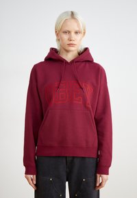 Μαρόν φούτερ με τσέπη μπροστά, κόκκινο κεντημένο κείμενο "OBEY", κουκούλα με κορδόνι και ριμπ μανσέτες. Το ύφασμα φαίνεται μαλακό και υφασμένο.