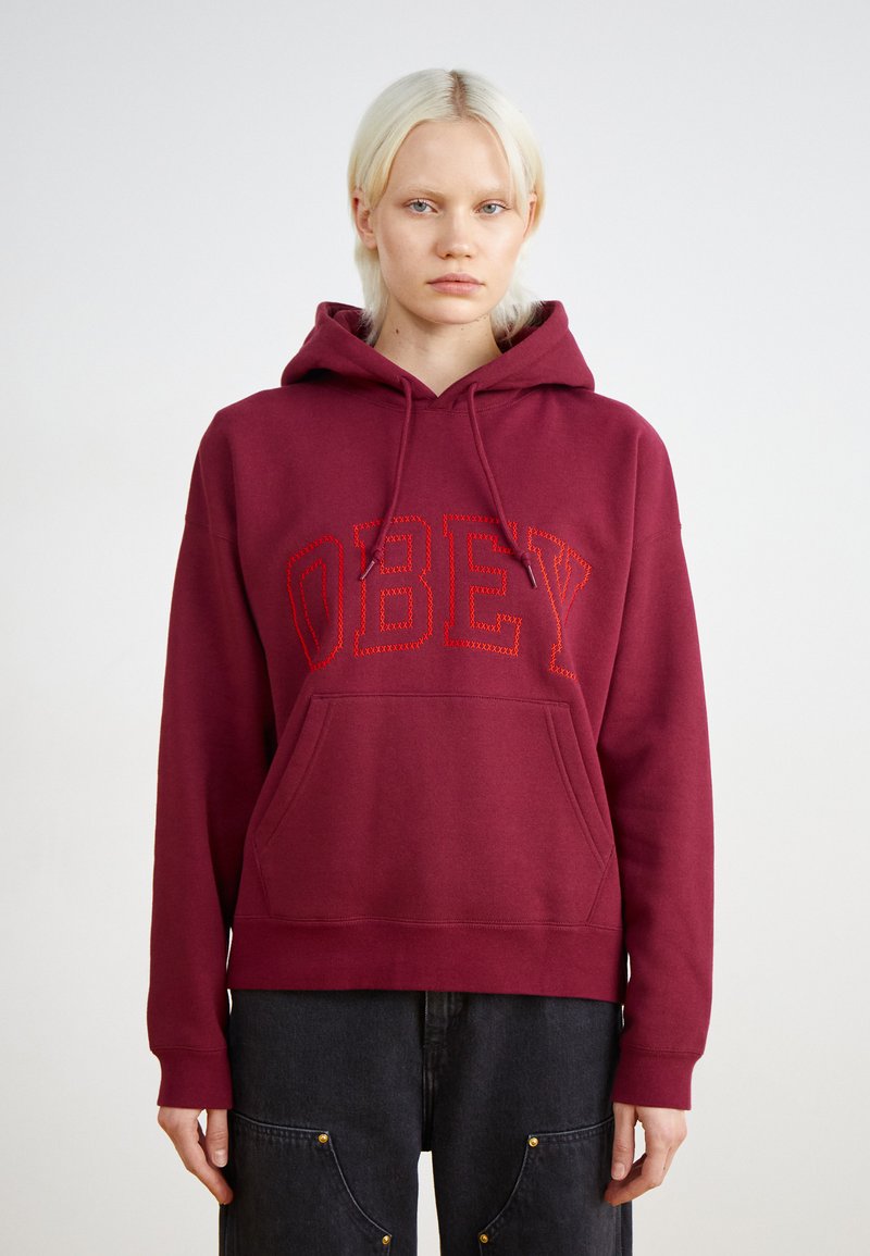 Μαρόν φούτερ με τσέπη μπροστά, κόκκινο κεντημένο κείμενο "OBEY", κουκούλα με κορδόνι και ριμπ μανσέτες. Το ύφασμα φαίνεται μαλακό και υφασμένο.