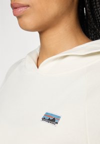Nærbilde av en person som har på seg en hvit hettegenser med et lite Patagonia-logo på brystet, som viser nakken og leppene i profil.