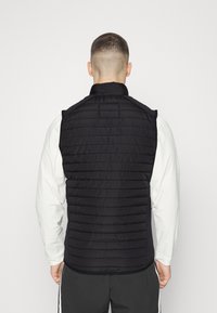 Schwarze Pufferweste mit einem gesteppten Muster, ausgestattet mit einem hohen Kragen und Schulternähten. Getragen über einem langen weißen Shirt.