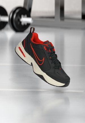 Schwarzer und roter Nike Air Monarch Sneaker mit weißer Sohle, schwebend über dem Boden des Fitnessraums, im Hintergrund ist eine Langhantel unscharf.