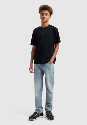 Tiener met een zwart T-shirt met kleine tekst, lichtblauwe uitgebleekte jeans en zwarte sneakers, staand met één hand in de zak tegen een effen achtergrond.