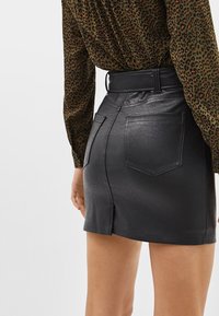 Černá mini sukně z umělé kůže s vysokým pasem, zadními kapsami a rozparkem. Stylizováno s vzorovanou košilí viditelnou nahoře.