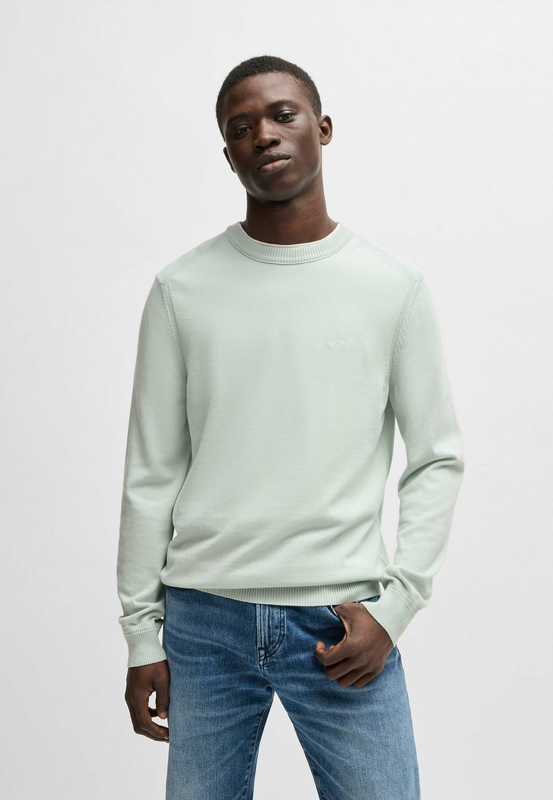 Maglione verde menta chiaro con scollo rotondo e polsini a costine. Presenta un sottile logo del marchio sul petto. Abbinato a jeans in denim blu.