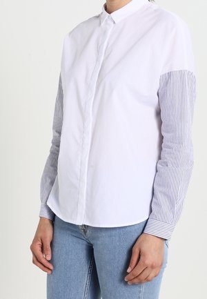 Persona con camisa blanca de botones con mangas largas a rayas azules y blancas y jeans azul claro, de pie contra un fondo liso.