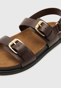 Bruine leren sandaal met twee banden, gouden gespsluiting en een tan suede binnenzool. Beschikt over een zwarte rubberen buitenzool voor grip.