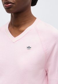 Detailní záběr osoby nosící světle růžový svetr s výstřihem do V a malým modro-bílým logem Adidas na hrudi.
