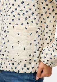 Gequilted jas in crème met marineblauwe bloempatronen. Voorzien van een ritssluiting aan de voorkant, elastische mouwen en een opgestikte zak. Zacht, tekstuur stof.