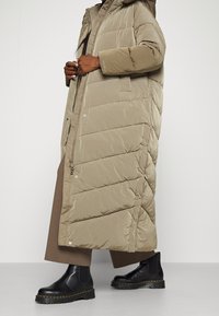 Abrigo acolchado beige con capucha, con secciones acolchadas y bolsillos laterales. Llevado sobre un conjunto marrón y combinado con botas negras hasta el tobillo.