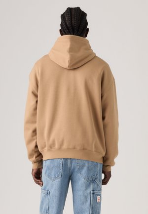 Person mit geflochtenem Haar, die einen beigen Hoodie und hellblaue Denim-Cargohosen trägt, mit dem Rücken zu einem schlichten hellen Hintergrund.