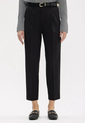 Zwarte op maat gemaakte enkellange broek met een zwarte riem en gouden gesp, gedragen met zwarte loafers en een top met lange mouwen en een patroon.