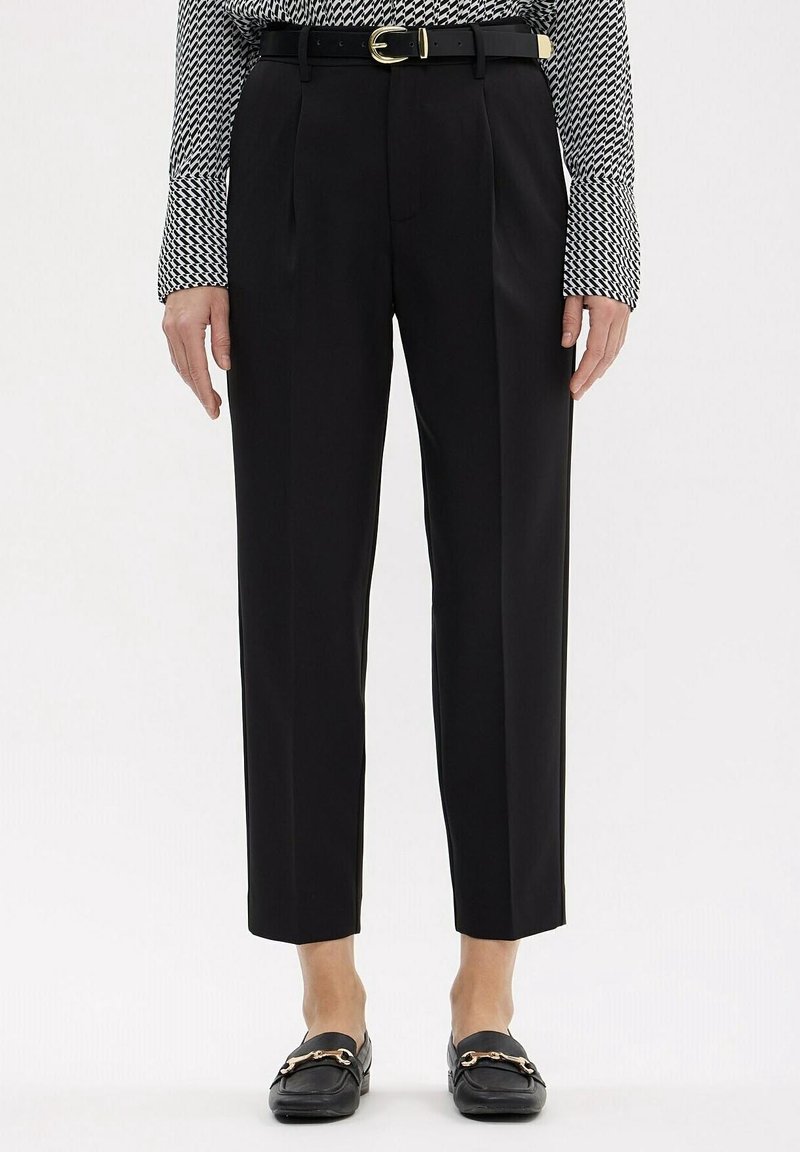 Pantalon noir ajusté longueur cheville avec une ceinture noire et une boucle dorée, porté avec des mocassins noirs et un haut à manches longues à motifs.