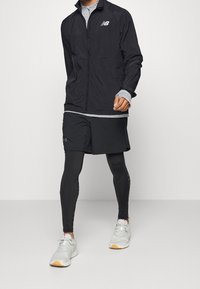 Homme marchant portant une veste noire New Balance, un short noir par-dessus des leggings noirs et des chaussures de sport blanches.