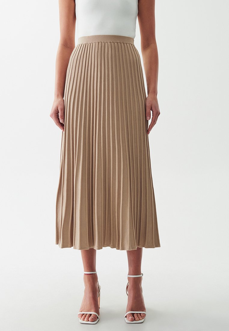 Gonna midi plissettata in maglia beige chiaro, con una vita alta e un design fluido. Indossata con tacchi bianchi con cinturini.