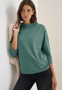 Top verde de manga larga con cuello tipo embudo, corte holgado y detalles de botones en los hombros. Combinado con jeans oscuros y una prenda beige.