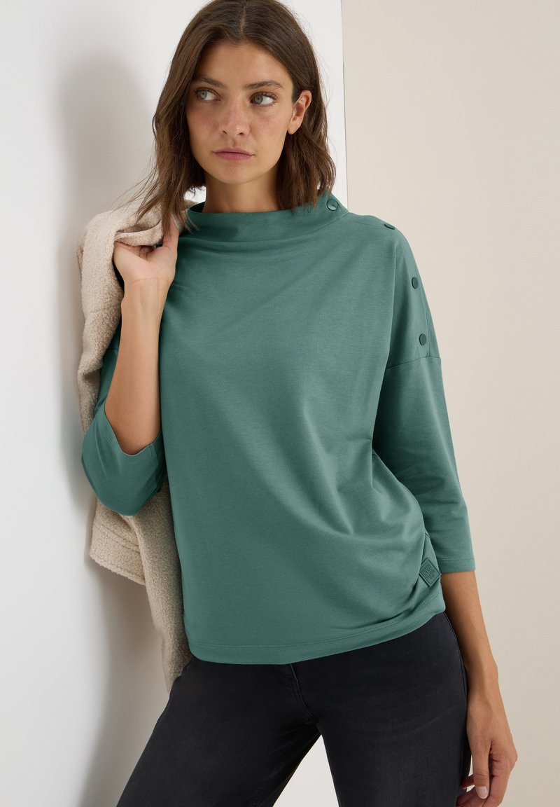 Top verde de manga larga con cuello tipo embudo, corte holgado y detalles de botones en los hombros. Combinado con jeans oscuros y una prenda beige.