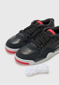 Schwarze Wildleder- und Leder-Sneaker mit roten Akzenten, ausgestattet mit einer grauen Sohle und schwarzen Schnürsenkeln. Enthält ein zusätzliches Paar weißer Schnürsenkel.