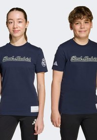 Námořnické modré tričká s nápisom "Real Madrid" bielym písaným písmom na hrudi. Rukávy zdobia športové logo a na spodnom leme je malá nášivka.