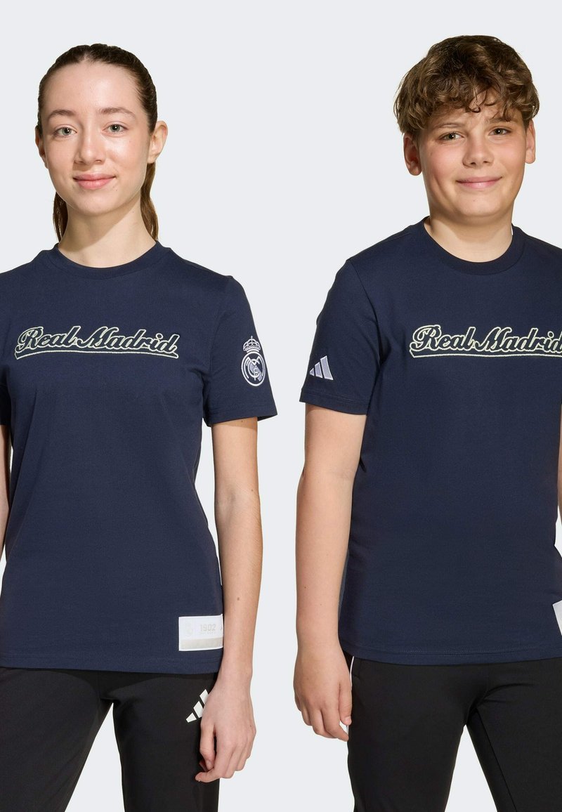 Námořnické modré tričká s nápisom "Real Madrid" bielym písaným písmom na hrudi. Rukávy zdobia športové logo a na spodnom leme je malá nášivka.