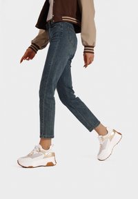 Sneaker bianche con dettagli dorati, materiali in tessuto e pelle, suola spessa e dettagli in rete. Indossate con jeans skinny blu e una giacca marrone.