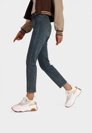 Sneaker bianche con dettagli dorati, materiali in tessuto e pelle, suola spessa e dettagli in rete. Indossate con jeans skinny blu e una giacca marrone.
