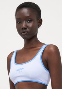 Fehér sportmelltartó világoskék részletekkel és lekerekített nyakkivágással. Középen kék logóval és vastag pántokkal rendelkezik. Simára végzett anyagtextúra.