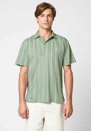 Giovane uomo con polo verde a maniche corte con strisce verticali bianche, in piedi contro uno sfondo chiaro uniforme, pantaloni bianchi visibili.