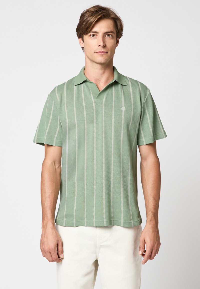 Jonge man draagt een groen polo met korte mouwen en witte verticale strepen, staand tegen een effen lichte achtergrond, witte broek zichtbaar.