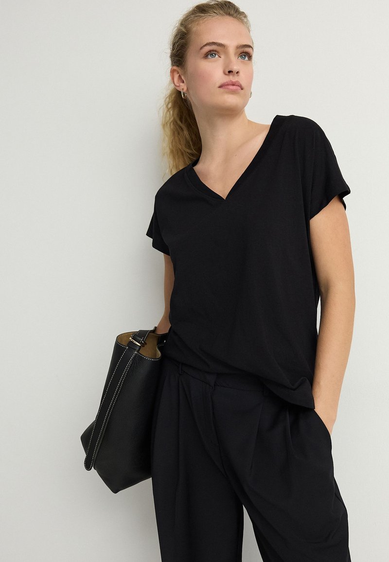 Jeune femme aux cheveux blonds attachés en queue de cheval, portant un haut noir à col en V et manches courtes ainsi qu’un pantalon noir, tenant un sac à main noir sur une épaule.