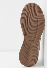 Zapato tipo slip-on de color gris claro con parte superior de tela texturizada y suela de goma marrón con ranuras horizontales. Nombre de la marca "BOBS" grabado en la suela.