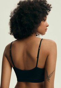 Czarny stanik o miękkiej, gładkiej fakturze, z regulowanymi ramiączkami i zaokrąglonym fasonem z tyłu. Na ramieniu znajduje się delikatny tatuaż.