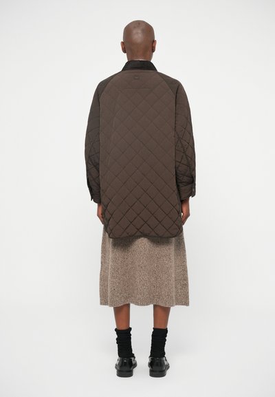 Casaco oversized castanho acolchoado com gola canelada, mangas longas e punhos abotoados, combinado com uma saia bege texturizada e botins pretos.