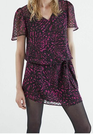 Femme portant une courte robe noire avec des manches transparentes et un motif floral rose, associée à des collants noirs opaques, debout devant un fond blanc.