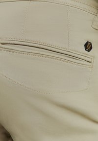 Details van beige broek met een gestructureerde stof, achterzak met decoratieve stiksels en een zwarte hexagonale accent met gouden lijnen.