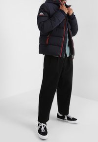 Marinblå jacka med vaddering och röda detaljer samt huva, matchad med en ljusblå skjorta, svarta byxor med avslappnad passform, och svarta sneakers med vita sulor.