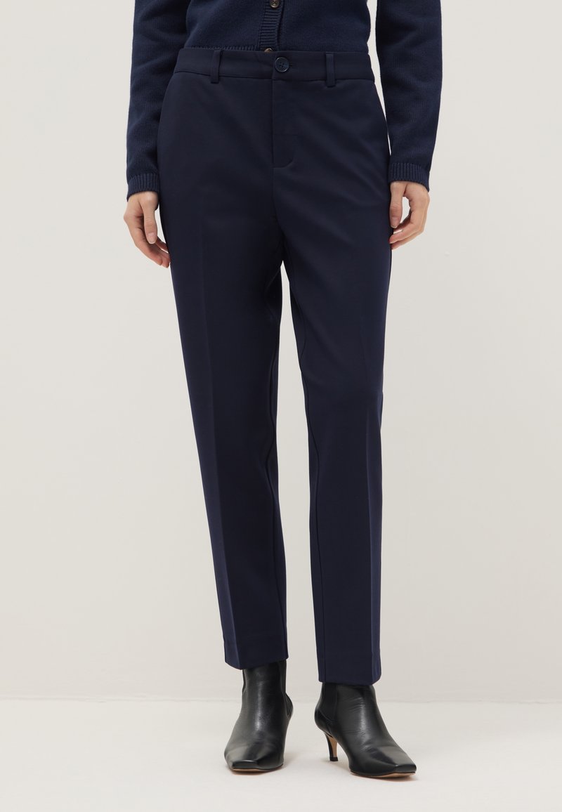 Frau trägt marineblaue gerade geschnittene Hose, marineblauen Cardigan und schwarze Stiefeletten mit Absatz, steht vor neutralem Hintergrund.