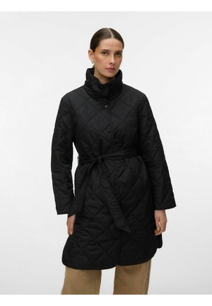 Kvinne iført en svart, quiltet lang frakk med høy krage og midjebelte, kombinert med beige bukser, stående mot en ensfarget hvit bakgrunn.