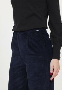 Pantalons en velours côtelé bleu marine avec des rayures verticales, dotés d'une taille plate, de poches latérales et d'une petite étiquette de marque près de la taille.