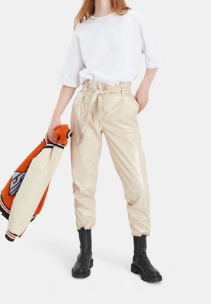 Woman wearing shiny beige tied paperbag pants, white oversized shirt, black boots, holding orange and cream varsity jacket in her left hand.