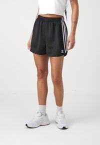 Černé sportovní šortky s bílými pruhy na bocích, elastickým pasem a logem na noze. Kombinovány s bílými teniskami a šedými ponožkami.