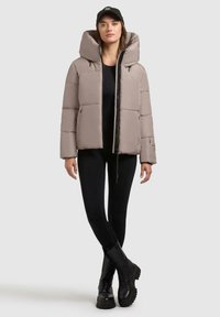 Beige pufferjakke med en stor hætte, frontlynlås og sidelommer, båret over en sort top og leggings. Sorte snørebånd ankle boots.