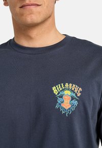 Tricou din bumbac de culoare bleumarin, prezentând o imprimare grafică colorată pe partea stângă a pieptului, cu textul "BILLABONG" și modele stilizate de valuri.