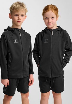 Twee kinderen in bijpassende zwarte zip-up hoodies met logo's en shorts. De stof lijkt glad, met zijzakken en een lichte ontwerp.