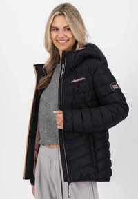 Lächelnde Frau in einer schwarzen gesteppte Jacke mit Kapuze, grauem cropped Pullover und hellgrauen Hosen vor einem schlichten weißen Hintergrund.